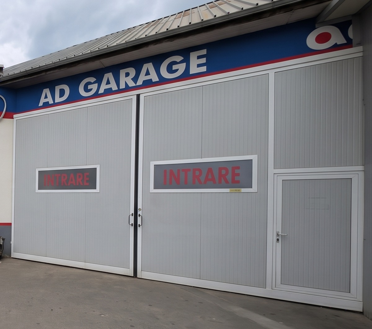 Die Fassade der Werkstatt: graue Tore mit der Aufschrift INTRARE (Einfahrt) unter einem blauen Band mit dem Schriftzug AD GARAGE